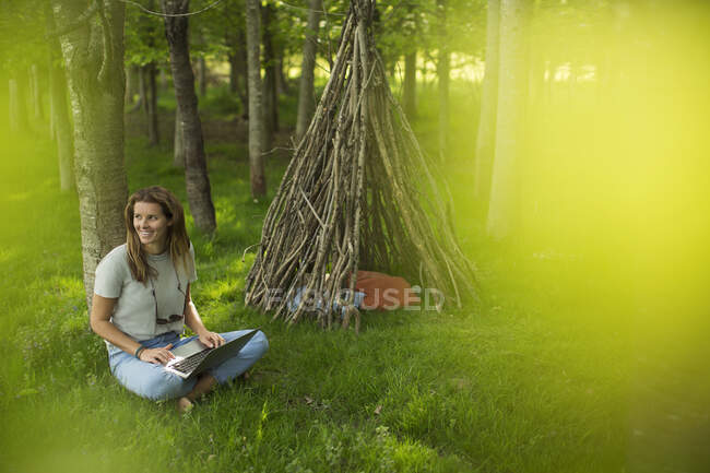 Улыбающаяся женщина с помощью ноутбука возле ветки вигвама в лесу — стоковое фото