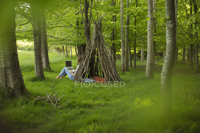 Young woman using laptop at branch teepee in woodland — Stock Photo