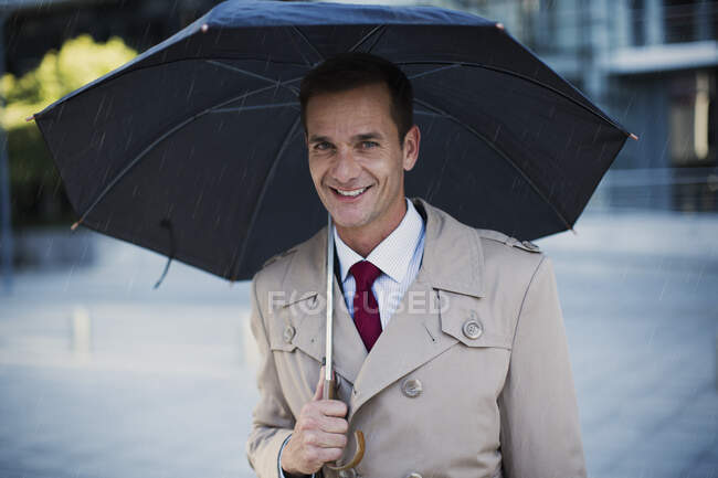 Hombre de negocios sonriente con gabardina bajo paraguas - foto de stock