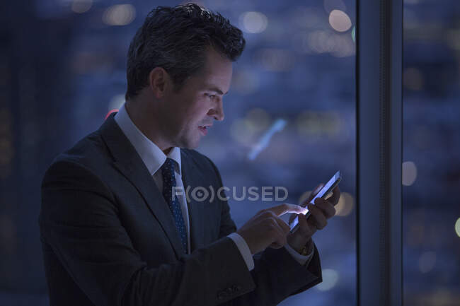 Mensajería de texto de hombre de negocios con teléfono celular en la ventana por la noche - foto de stock