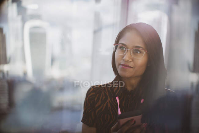 Задумчивая деловая женщина со смартфоном в солнечном окне — стоковое фото