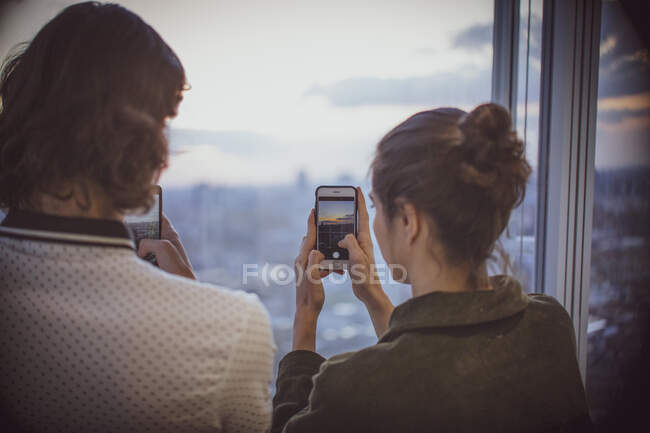 Pareja joven con teléfonos inteligentes fotografiando la puesta de sol en la ventana de rascacielos - foto de stock