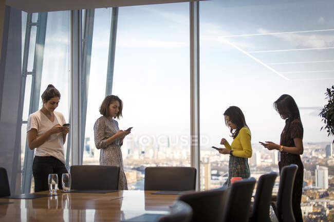Businesswomen utilizando teléfonos inteligentes en la ventana de la oficina de rascacielos - foto de stock