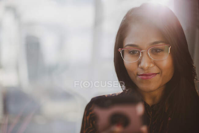 Empresária sorridente usando telefone inteligente na janela ensolarada — Fotografia de Stock