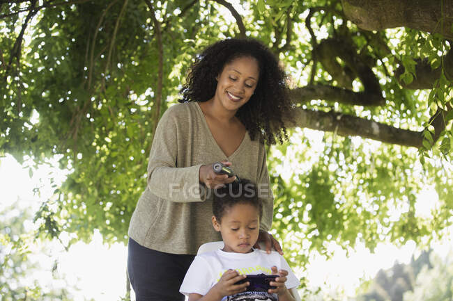 Mãe aparando o cabelo do filho debaixo da árvore no quintal de verão — Fotografia de Stock