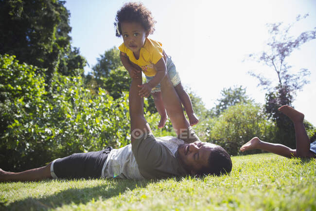 Pai levantando bonito criança filha sobrecarga na grama ensolarada verão — Fotografia de Stock