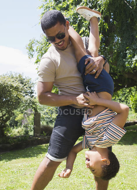 Игривый отец держит сына вверх ногами на солнечном летнем заднем дворе — стоковое фото