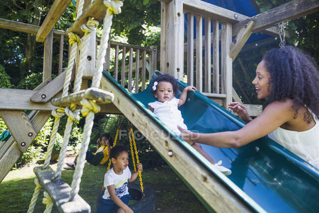 Mother watching cute toddler daughter on playground slide in backyard — Stock Photo