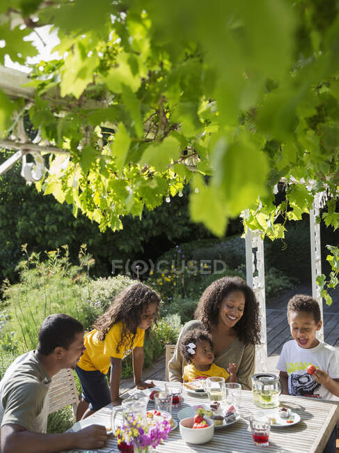 Familia feliz disfrutando del almuerzo en la soleada mesa de jardín de verano - foto de stock