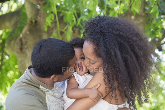 Affectionate parents kissing toddler daughter below tree — Stock Photo