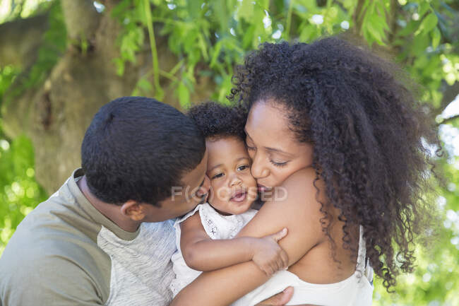 Affectionate parents kissing toddler daughter under tree — Stock Photo