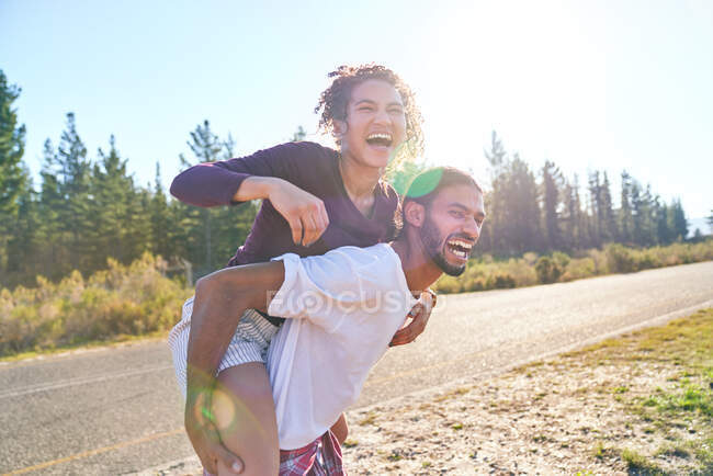 Happy playful young couple piggybacking on sunny summer roadside — Stock Photo