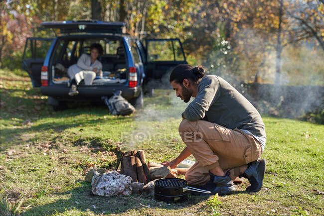 Man preparing campfire outside car in sunny autumn field — Stock Photo