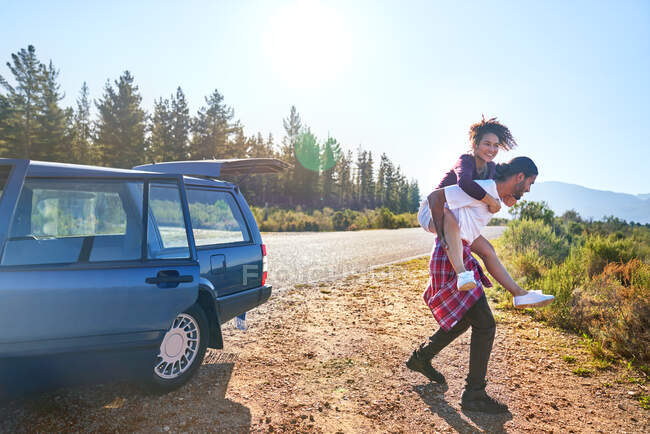 Carefree young couple piggybacking outside car at sunny roadside — Stock Photo