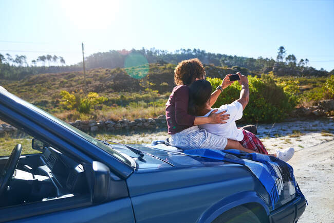 Carefree young couple taking selfie on hood of car at sunny roadside — Stock Photo