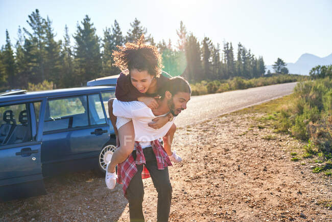 Playful young couple piggybacking outside car at sunny remote roadside — Stock Photo