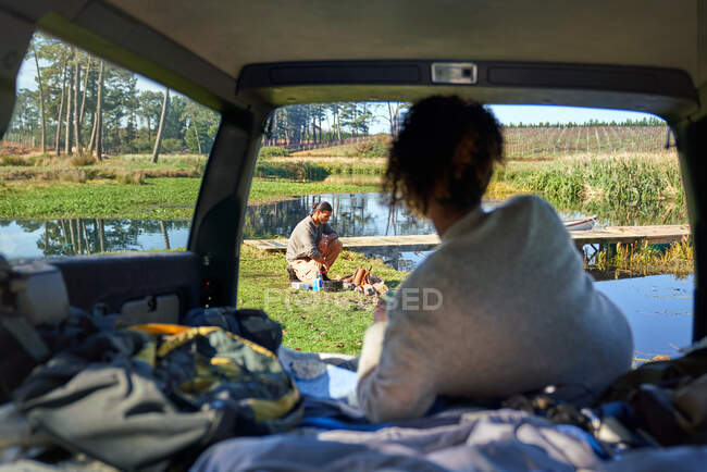 Jeune femme en voiture regarder copain préparer feu de camp — Photo de stock