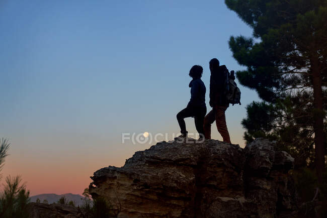 Silhueta jovem caminhante casal desfrutando vista da lua da rocha ao entardecer — Fotografia de Stock