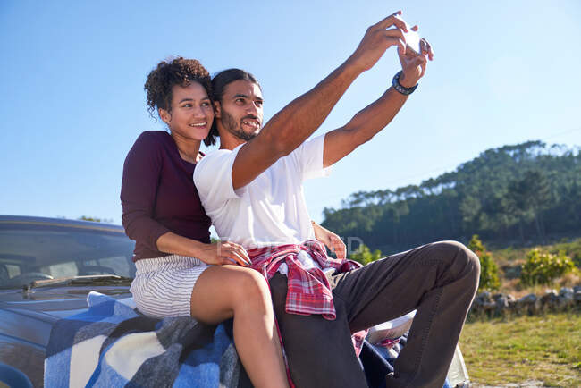 Heureux jeune couple prenant selfie sur le capot de la voiture au bord de la route ensoleillée — Photo de stock