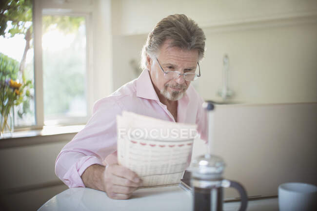 Hombre mayor leyendo el periódico en el portátil en la mesa de la cocina de la mañana - foto de stock
