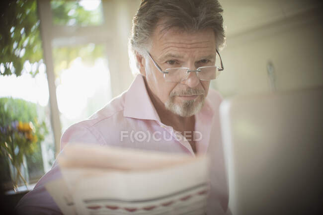Senior man reading newspaper at laptop in sunny morning kitchen — Stock Photo