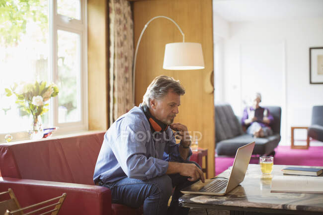 Homme âgé avec écouteurs travaillant à l'ordinateur portable sur le canapé du salon — Photo de stock