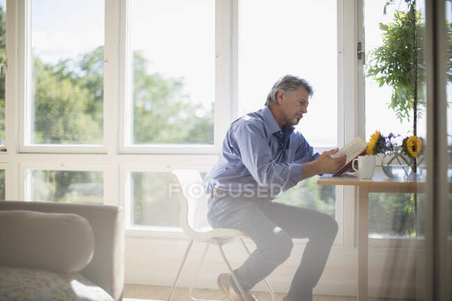 Hombre mayor leyendo libro en la mesa de comedor por ventana soleada - foto de stock