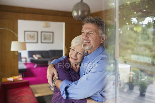 Glückliches Senioren-Paar umarmt sich am sonnigen Wohnzimmerfenster — Stockfoto