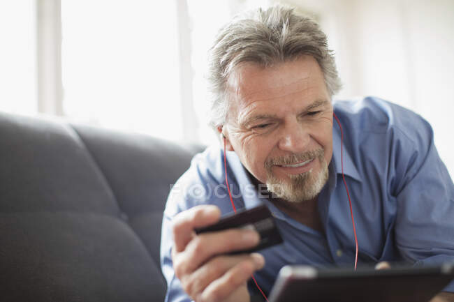 Homme âgé avec casque et carte de crédit en utilisant une tablette numérique sur le canapé — Photo de stock