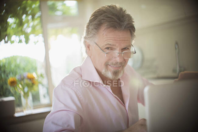 Retrato sorrindo homem sênior trabalhando no laptop na cozinha ensolarada — Fotografia de Stock