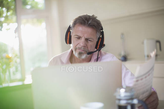 Старший чоловік з навушниками та газетами, що працюють на ноутбуці на кухні — стокове фото
