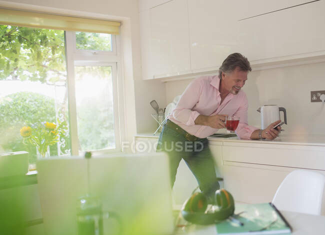 Senior man drinking tea and using smart phone in sunny kitchen — Stock Photo