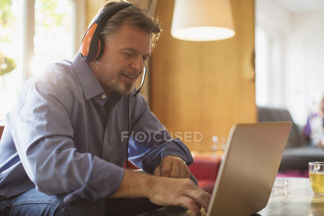 Старший чоловік з навушниками використовує ноутбук на дивані у вітальні — стокове фото