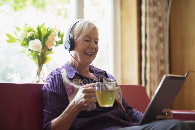 Donna anziana felice con cuffie e tablet digitale bere tè — Foto stock