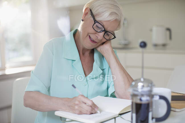 Mujer mayor escribiendo en el diario en la cocina de la mañana - foto de stock