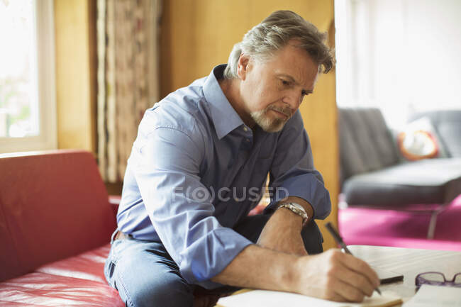 Senior man writing in notebook at living room coffee table — Stock Photo