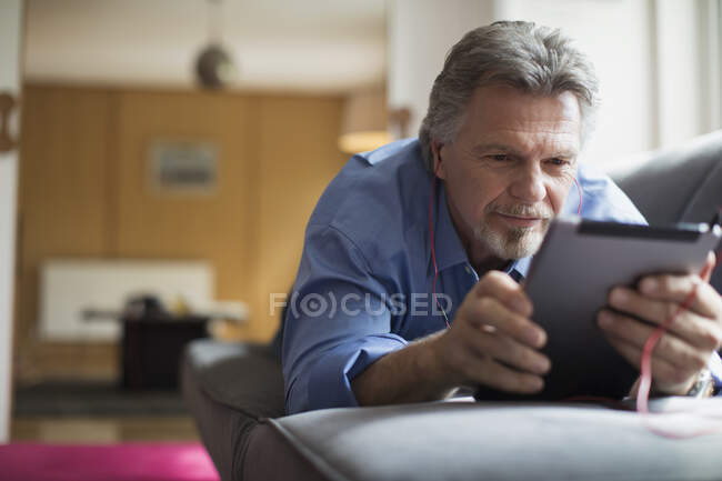 Homem idoso usando fones de ouvido e tablet digital no sofá da sala de estar — Fotografia de Stock