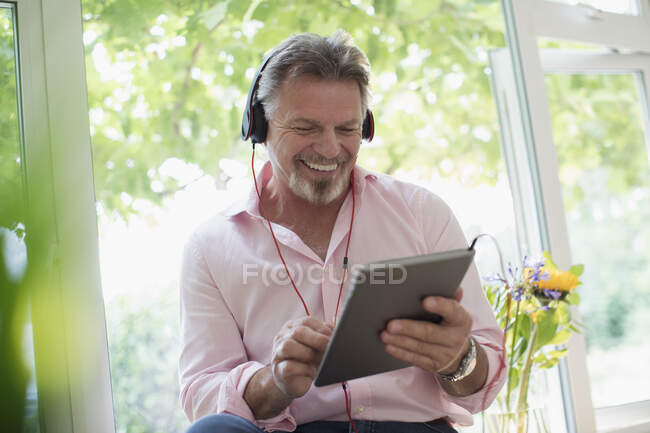 Homem sênior feliz usando fones de ouvido e tablet digital na janela — Fotografia de Stock