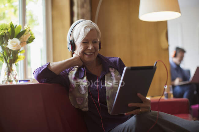 Щаслива старша жінка, яка користується навушниками і цифровим планшетом на дивані. — стокове фото