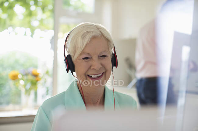 Mujer mayor feliz con auriculares usando el ordenador portátil en la cocina - foto de stock