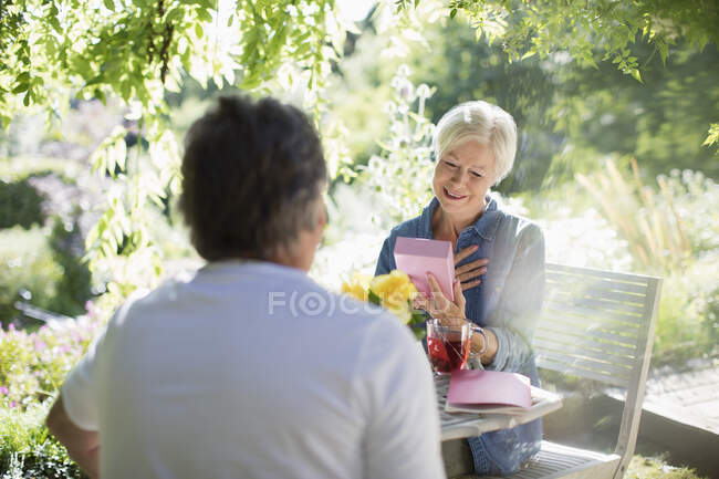 Счастливая пожилая женщина открывает подарок от мужа на солнечном летнем дворике — стоковое фото
