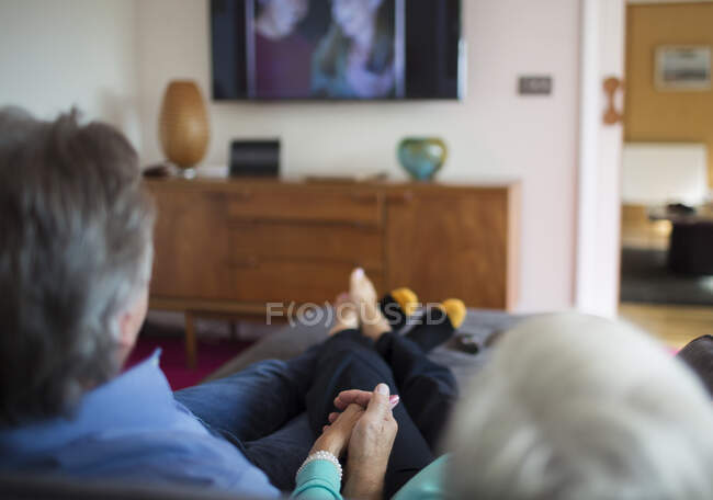 Senior couple holding hands and watching TV on living room sofa — Stock Photo