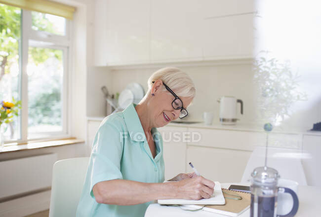 Donna anziana che scrive in diario al tavolo della cucina del mattino — Foto stock