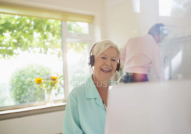 Happy senior woman with headphones video chatting at laptop in kitchen — Stock Photo