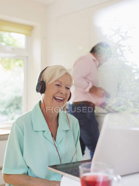 Heureuse femme âgée avec écouteurs vidéo bavardage à l'ordinateur portable dans la cuisine — Photo de stock