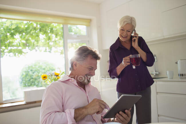 Счастливая пожилая пара с помощью цифрового планшета и смартфона на кухне — стоковое фото