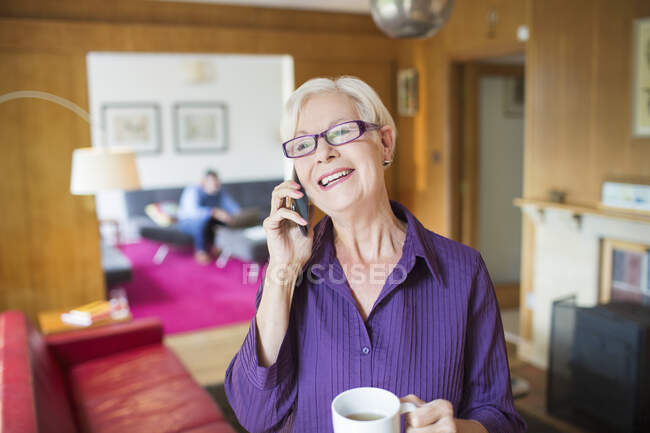 Mulher sênior feliz com café falando no telefone inteligente na sala de estar — Fotografia de Stock