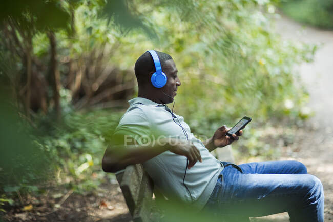 Мужчина отдыхает на скамейке в парке с наушниками и смартфоном — стоковое фото