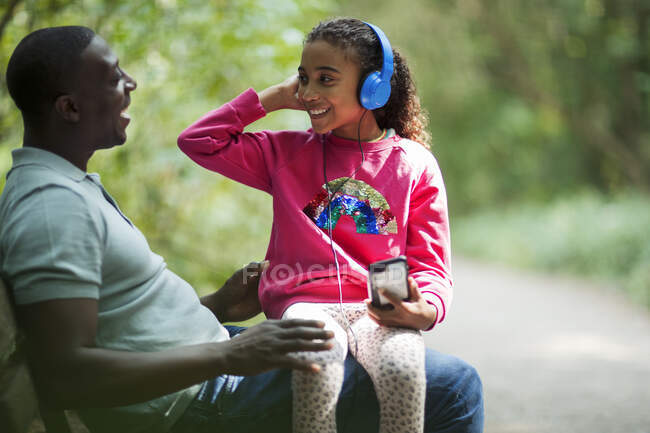 Feliz padre e hija sentados en el banco del parque con auriculares - foto de stock