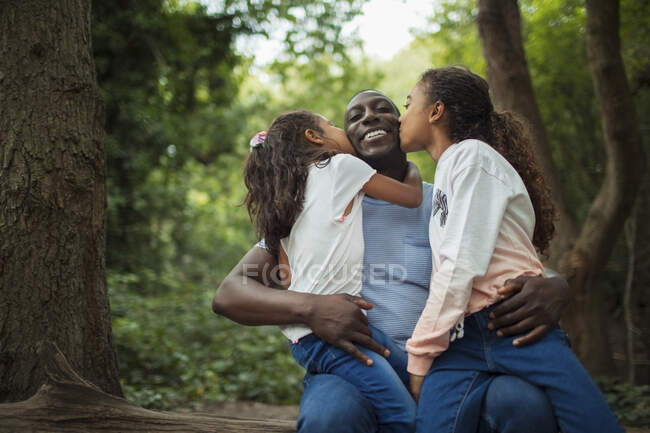 Happy affectionate daughters kissing father below trees in woods — Stock Photo